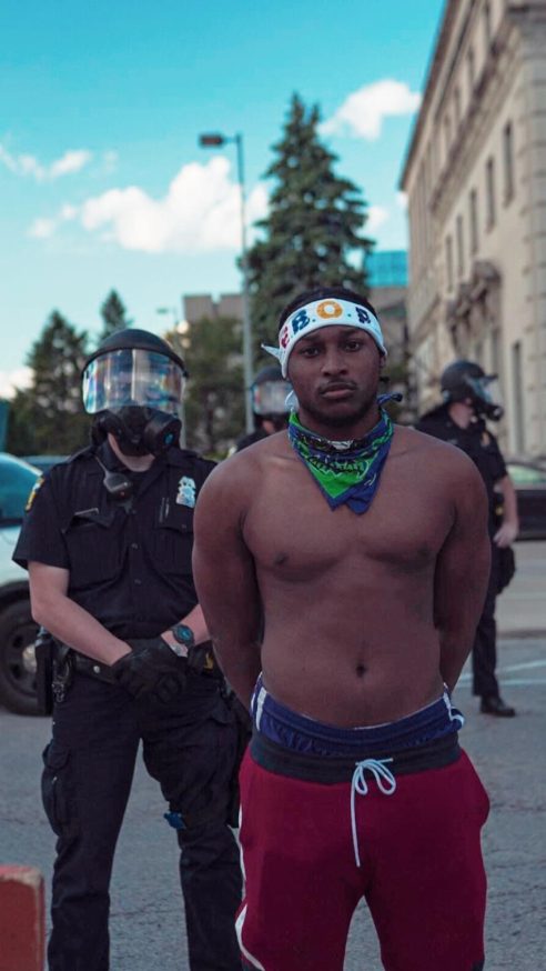 Ebop standing calm with his hands behind his back in front of police in riot gear
